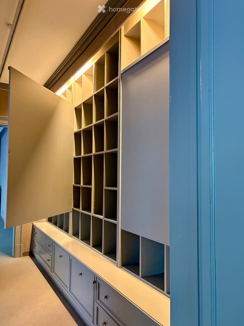An empty room with built-in storage units featuring multiple shelves and drawers, illuminated by soft lighting. The walls are painted in a light blue color, and the floor is carpeted.