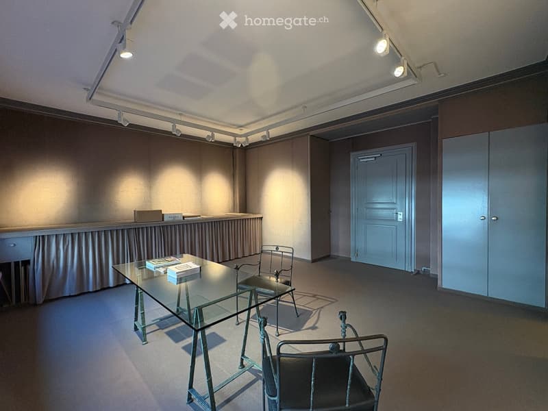 A modern, minimalist room with a glass table, two metal chairs, and a small counter with books and a box. The room features soft lighting from track lights and has a neutral color palette with carpeted flooring.