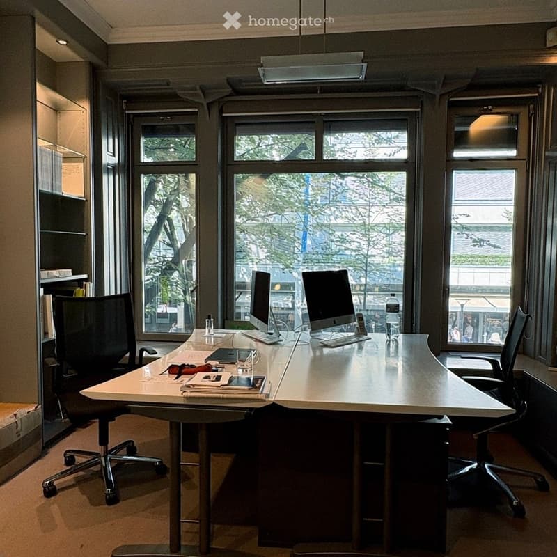 Modern office space with large windows, a long desk, two computer setups, office chairs, and various office supplies.