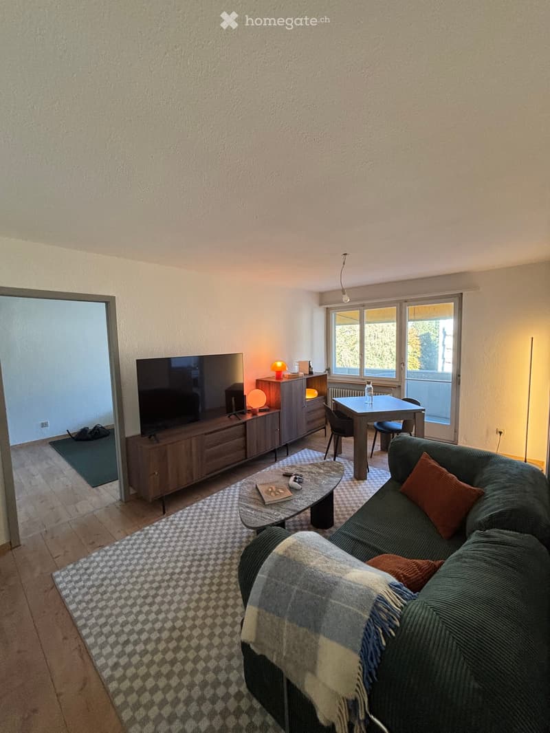 Living room with a green sofa, coffee table, television, sideboard, dining table, and chairs. The room features wooden flooring, a patterned rug, and large windows with a view.