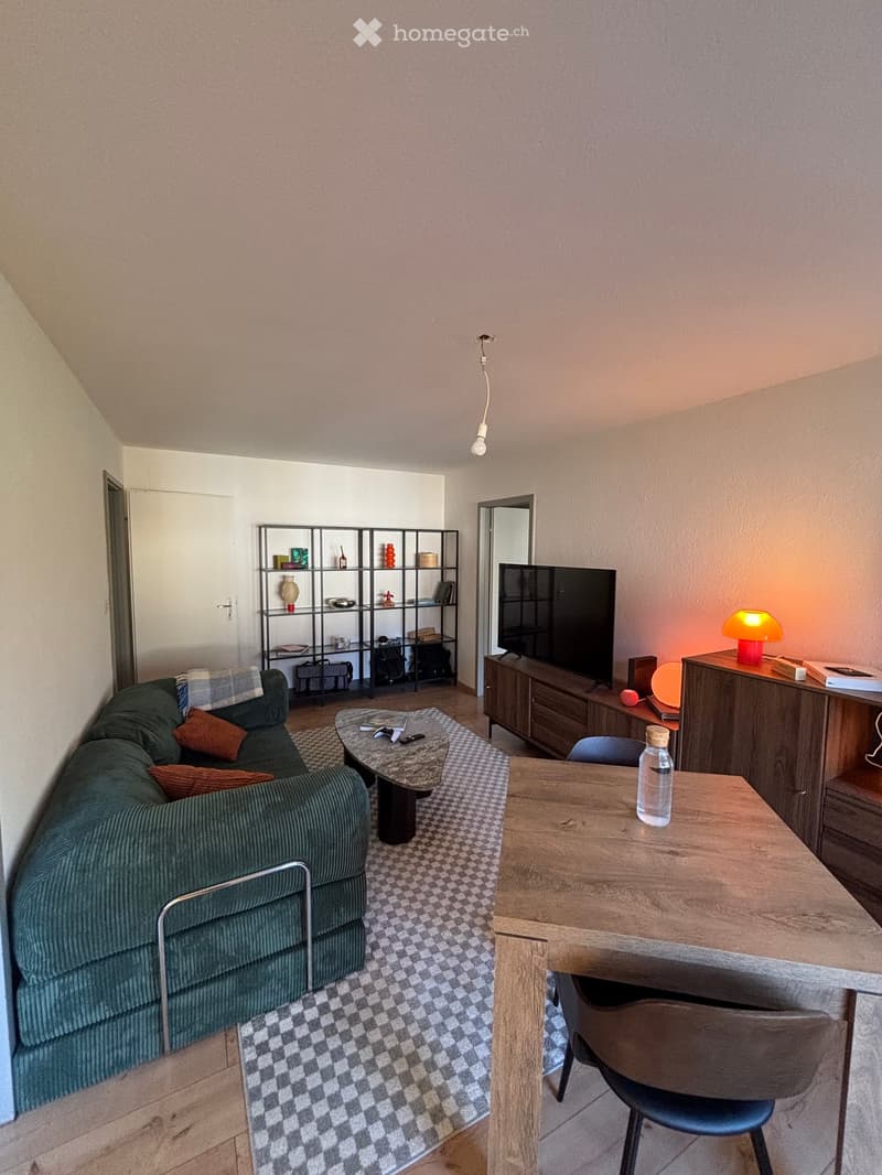 A modern living room with a green sofa, wooden coffee table, TV, and a small dining area with a wooden table and chair.