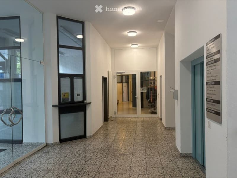 Clean, modern hallway with tiled flooring, recessed lighting, glass doors, and a visible signboard on the right side.