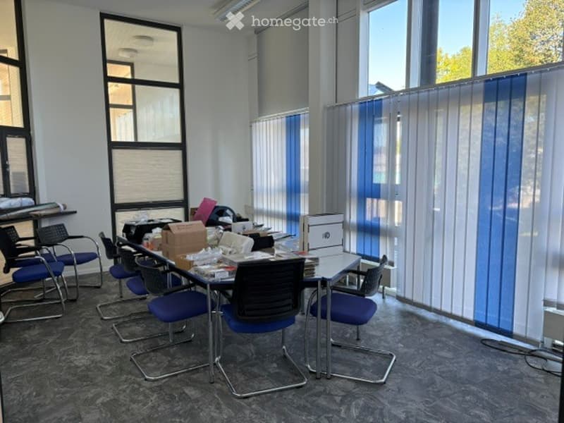 Office space with a table, chairs, and a screen, surrounded by large windows with blinds.