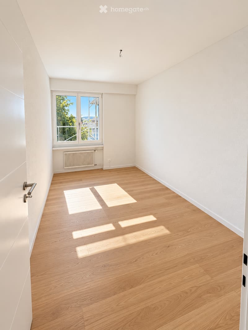Empty room with light wooden flooring, white walls, and a large window with a view of greenery. The room features a radiator below the window and a door on the left side.