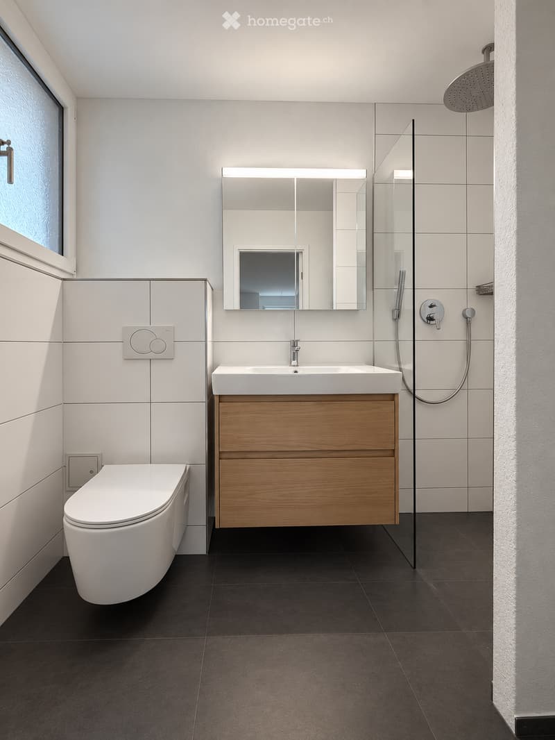 Modern bathroom with white tiles, dark floor, wooden vanity, sink, toilet, mirror, and glass-enclosed shower area.
