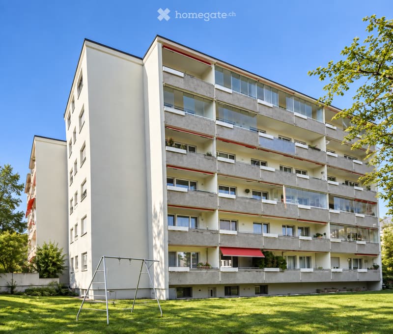 Multi-story residential building with balconies, large windows, and a playground in the foreground.