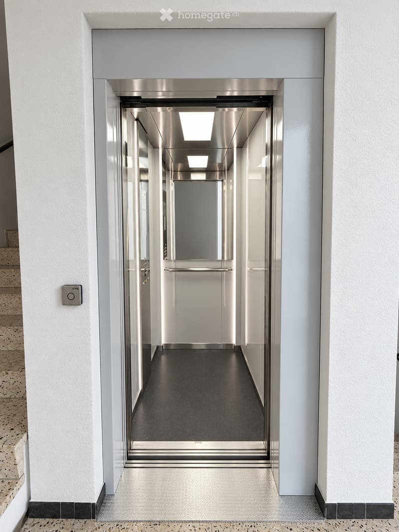 Modern hallway with white walls, dark carpeted floor, and an elevator with stainless steel doors. The hallway features a polished concrete floor with a black and white tiled border and a staircase visible on the left side.