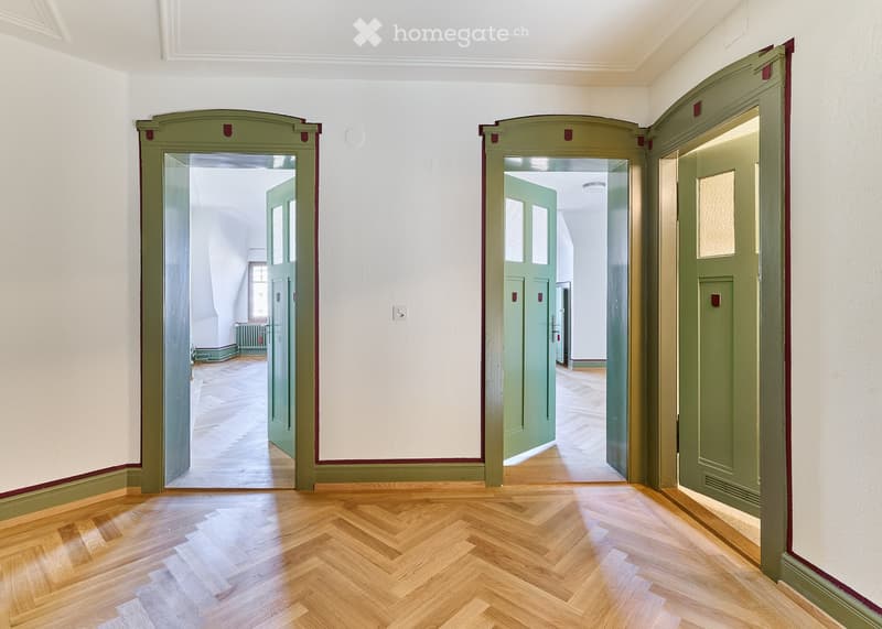 Empty hallway with wooden herringbone flooring, white walls, and green double doors leading to other rooms.