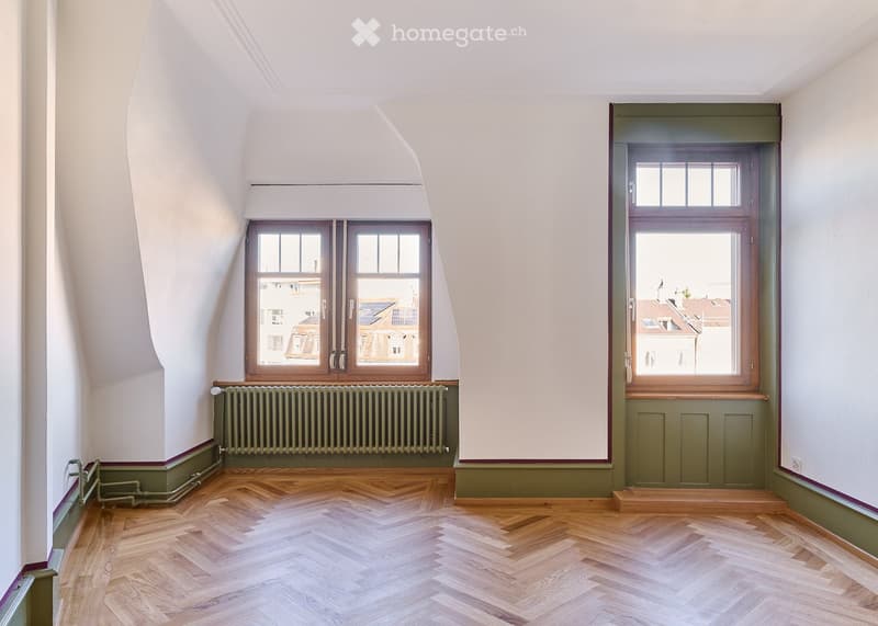 Empty attic room with wooden herringbone flooring, white walls, and large windows offering city views. Green radiator and baseboards visible.