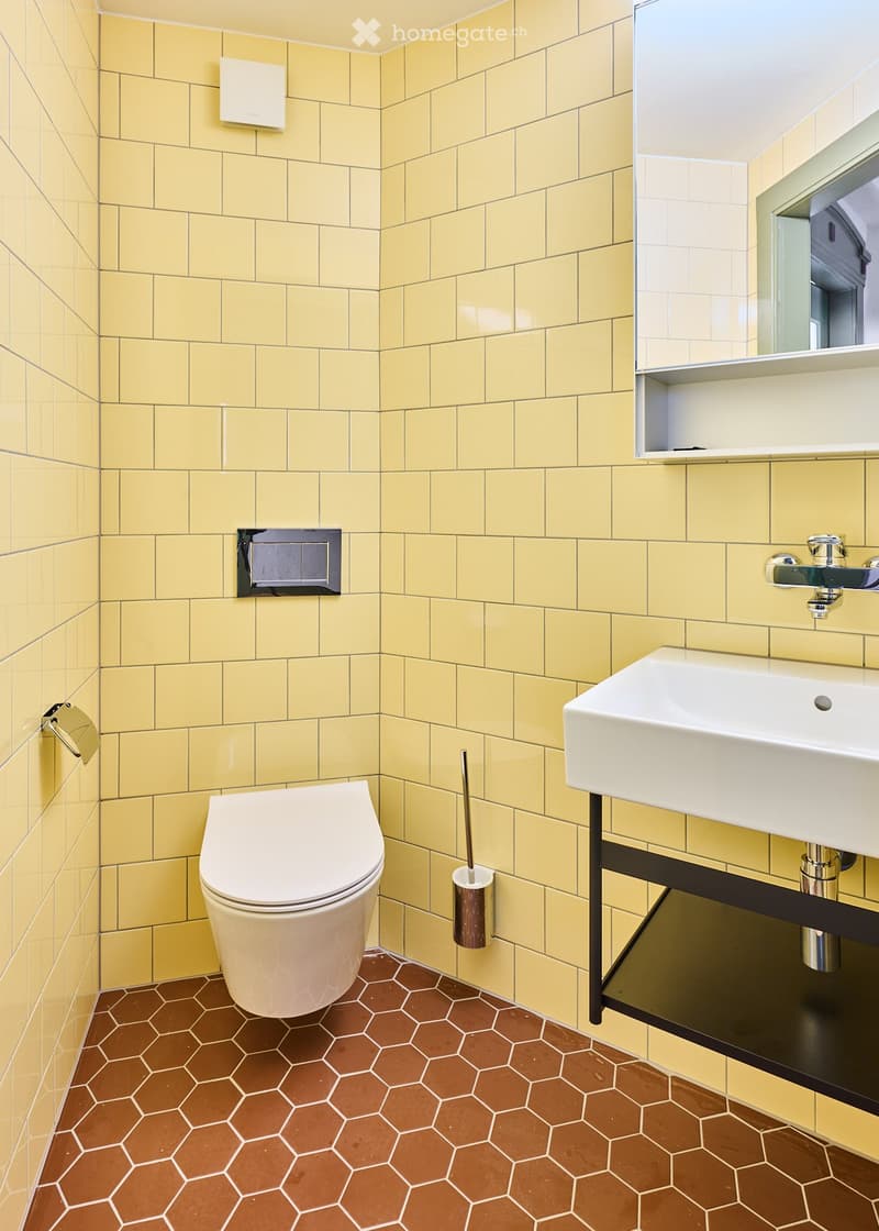 Modern bathroom with yellow tiled walls, brown hexagonal floor tiles, white toilet, white sink with chrome faucet, and a mirror above the sink.