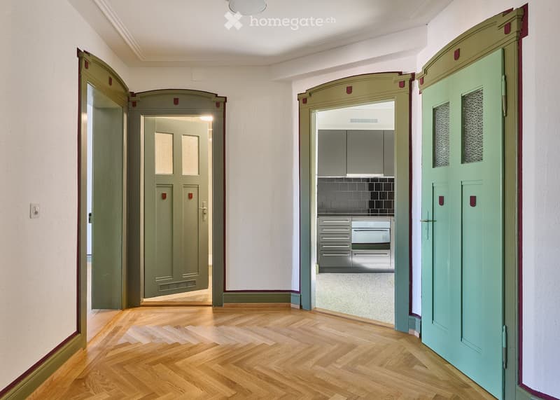 A hallway with wooden flooring, white walls, and several doors leading to different rooms, including a kitchen with stainless steel cabinets and drawers.