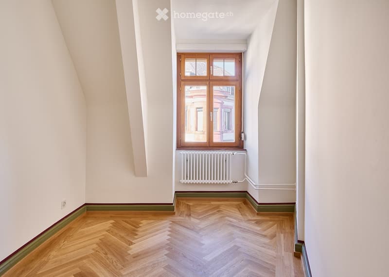 Empty room with wooden herringbone flooring, white walls, and a large window with wooden frame. A radiator is mounted below the window.
