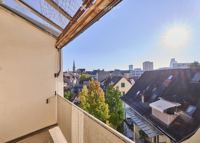 Balcony overlooking a cityscape with rooftops, trees, and distant buildings under a clear blue sky.
