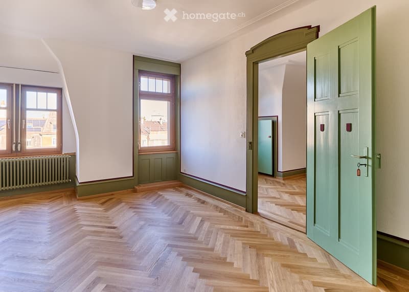 Empty room with wooden herringbone flooring, white walls, and green trim. Two windows with wooden frames and a radiator beneath one window. A green door is open to an adjacent space.