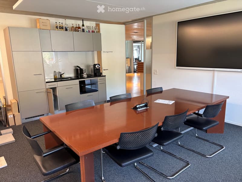 A modern conference room with a large wooden table, black office chairs, a flat-screen TV, and a small kitchenette area with cabinets, sink, and coffee machine.