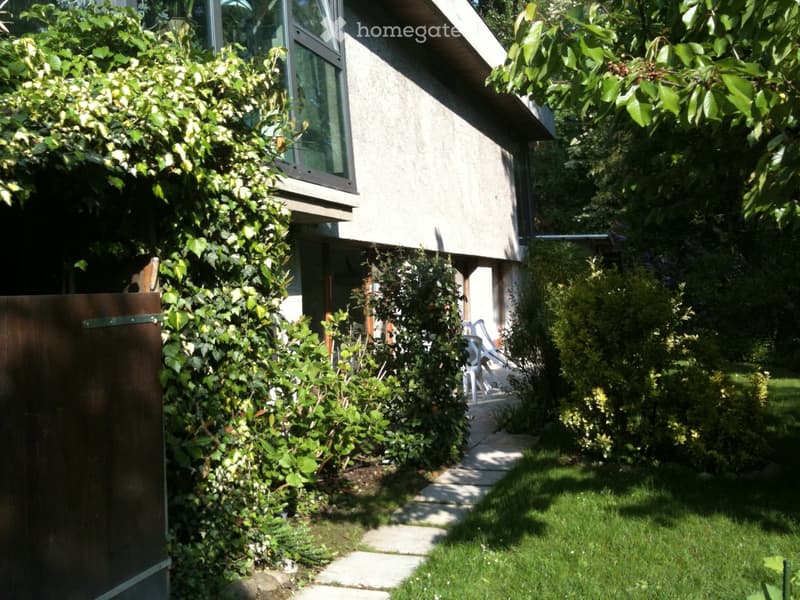 A modern house with a well-maintained garden, featuring a stone pathway, lush greenery, and a small seating area with white chairs near the entrance.