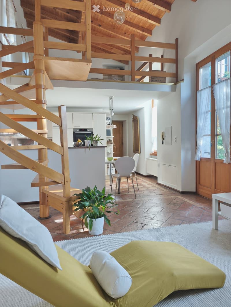 Open-plan living and dining area with a green sofa, white chair, dining table, kitchen area with oven, and a staircase leading to an upper level.