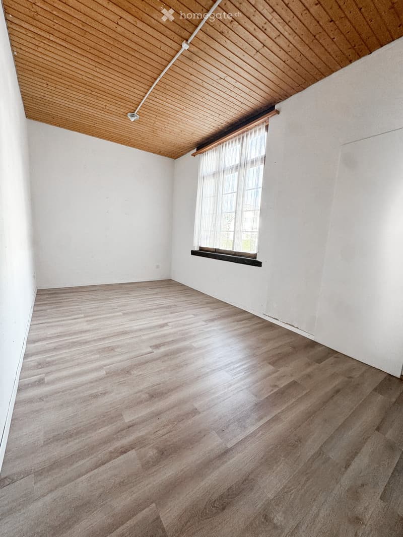 Empty room with wooden flooring and white walls, featuring a slanted wooden ceiling and a window with sheer curtains.
