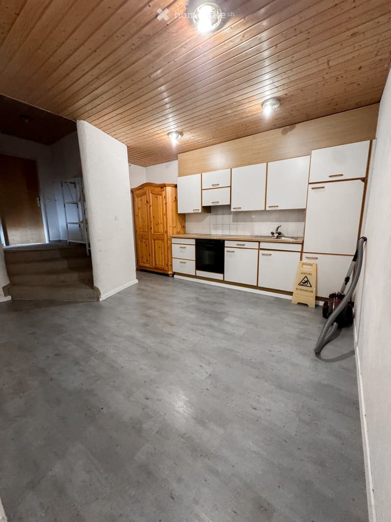 Empty kitchen with white cabinets, a wooden wardrobe, and a concrete floor. No furniture or appliances are present.