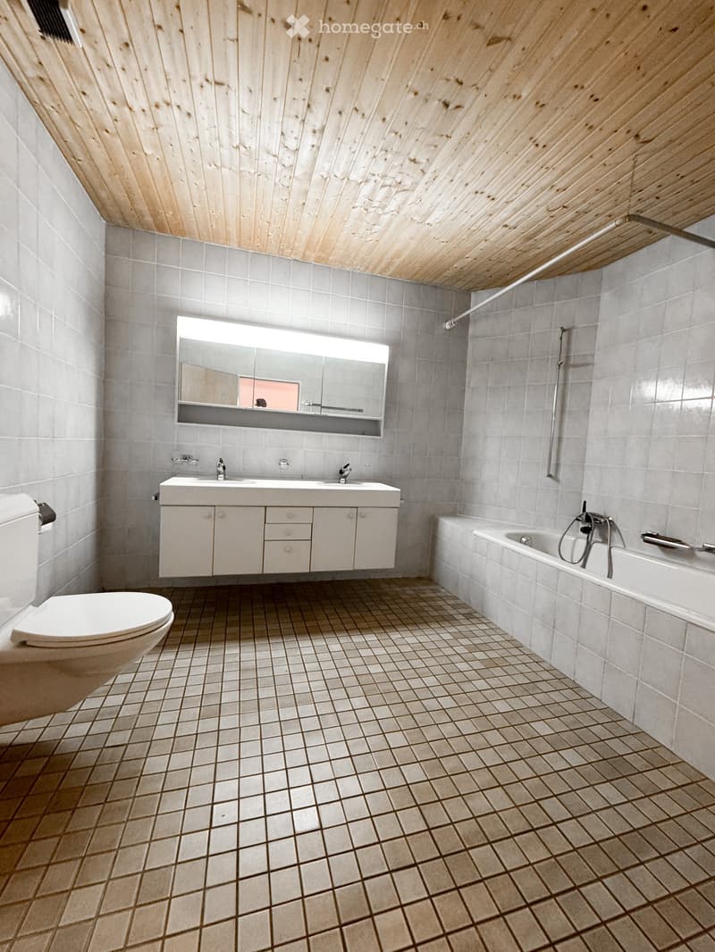 Modern bathroom with grey tiles, white sink, bathtub, toilet, and large mirror above the sink.