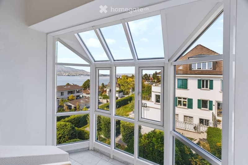 Bright room with large windows offering scenic views of a lake and surrounding houses, featuring a white sofa and tiled floor.