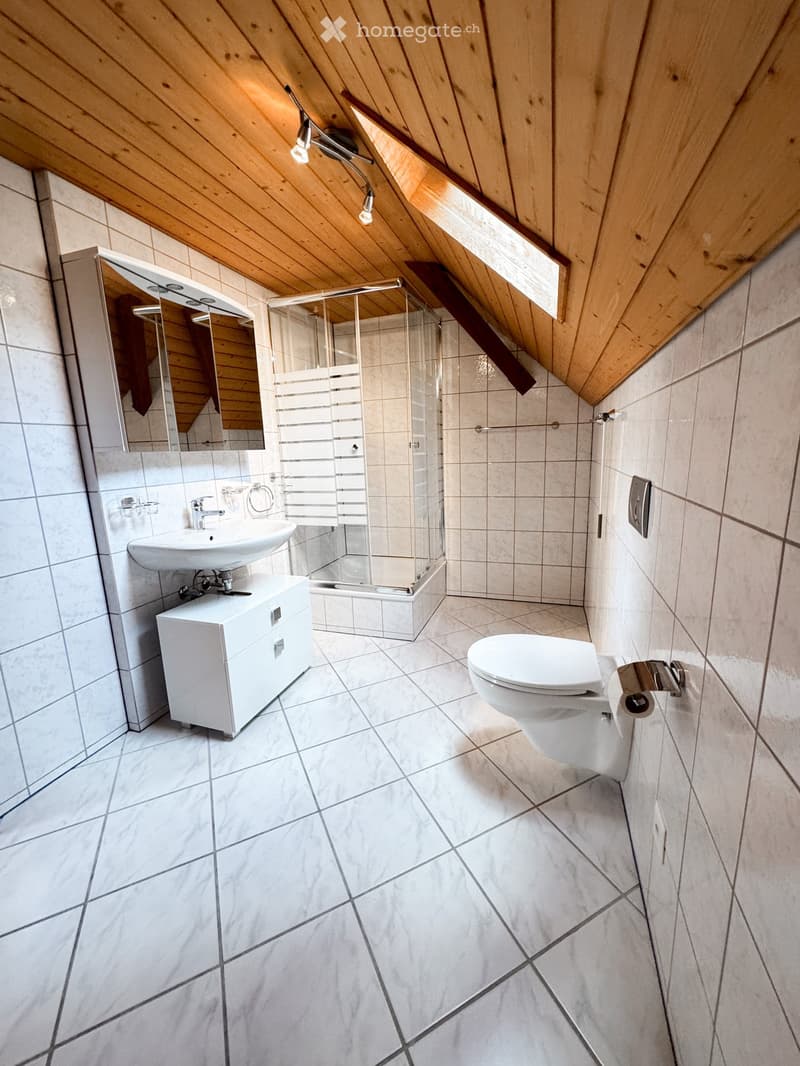 Modern bathroom with white tiles, a sink with a cabinet, a toilet, a glass-enclosed shower, and a mirror above the sink. The ceiling has wooden slats and built-in lighting.
