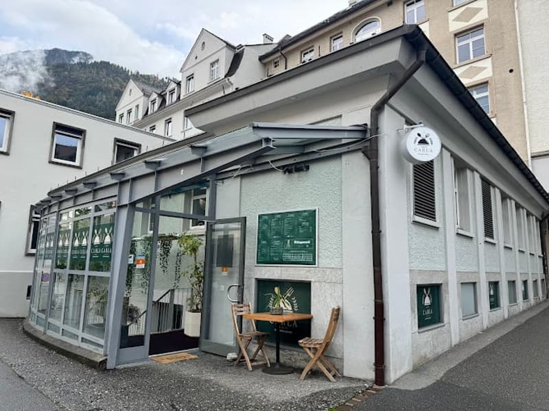 Small outdoor seating area of a café with a table, two chairs, and a potted plant. The café has a modern glass enclosure and a menu board visible on the wall.