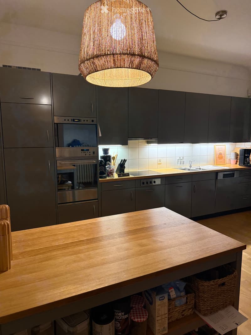 Modern kitchen with dark grey cabinets, stainless steel appliances, a wooden island, and a hanging pendant light. Visible items include a coffee maker, knife block, and various kitchen utensils.
