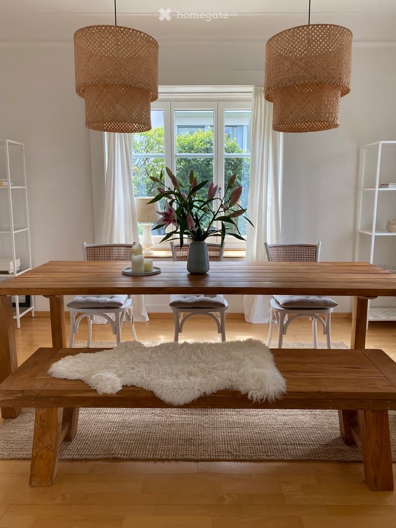 Bright dining area with wooden table, chairs, bench, vase with flowers, and hanging woven lamps.
