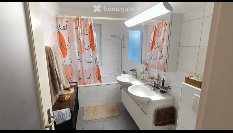 Modern bathroom with white tiles, double sink vanity, bathtub, and large mirror. Includes towels, toiletries, and a small shelf.
