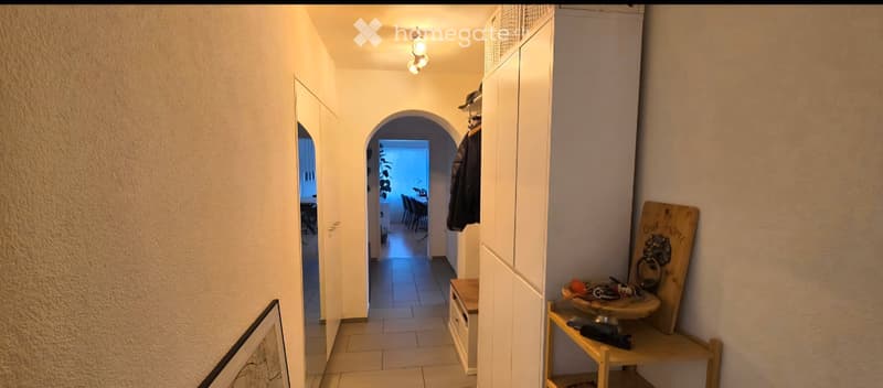 A narrow hallway with beige walls, tiled floor, and recessed lighting. A coat rack with jackets, a small wooden shelf with decorative items, and an arched doorway leading to another room are visible.