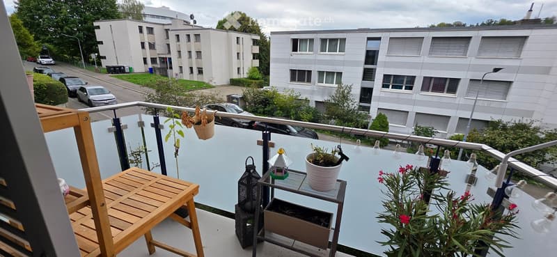 Balcony with wooden table, hanging plants, and string lights, overlooking a residential area with parked cars and buildings.