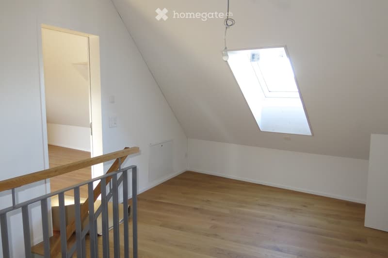 Bright, empty hallway with wooden flooring, white walls, and a skylight window at the far end. A staircase with a wooden railing is visible on the left side.