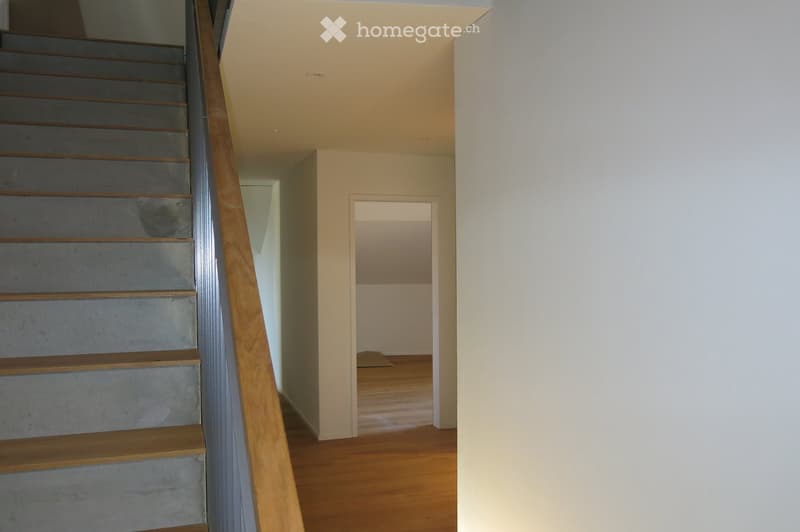 A modern staircase with wooden handrails and grey concrete steps leads to an open hallway with wooden flooring and white walls.