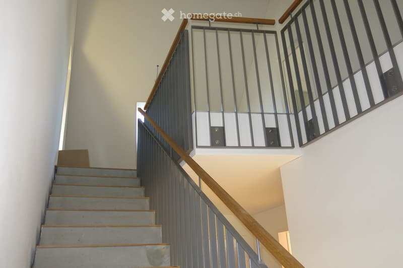 Modern staircase with grey concrete steps, wooden handrails, and metal balustrade featuring glass panels. The walls are painted in a light neutral tone, and the lighting is bright and even.