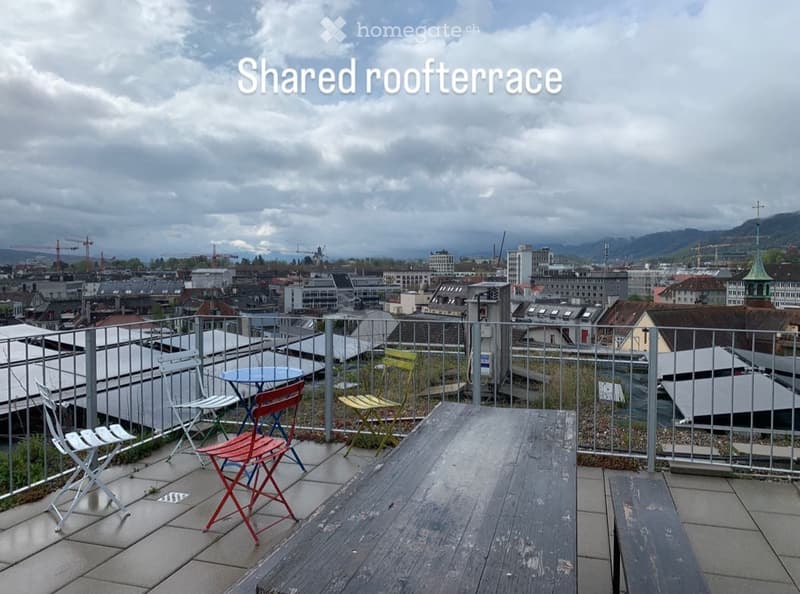 Shared rooftop terrace with a cityscape view, featuring a table and chairs, a wooden bench, and a metal railing.