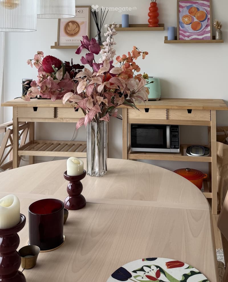 A modern kitchen with a wooden dining table, vase of flowers, candles, microwave, and teapot on shelves.