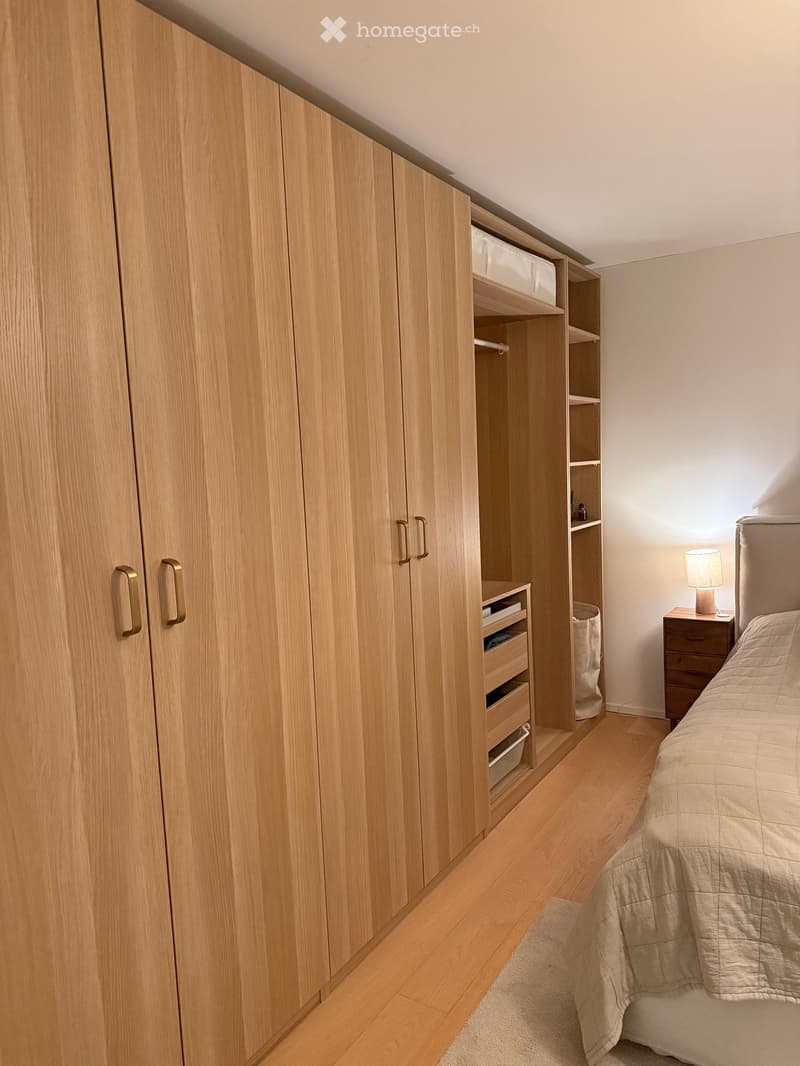 Bedroom with a neatly made bed, a wooden wardrobe with built-in storage, a nightstand with a lamp, and a wooden floor.