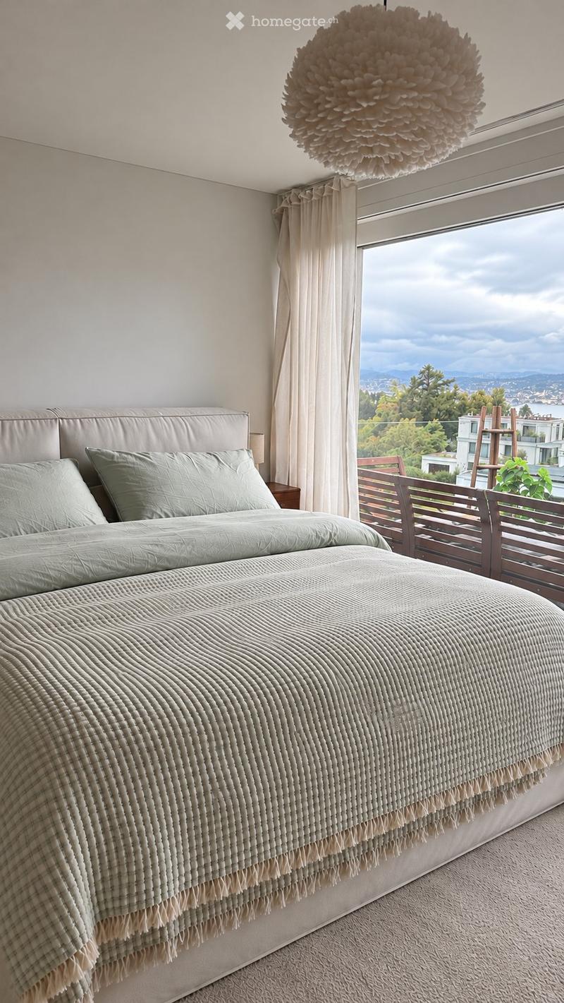 Modern bedroom with a large bed, nightstand, and a view of the ocean through large windows. The room features a pendant light and light-colored bedding.
