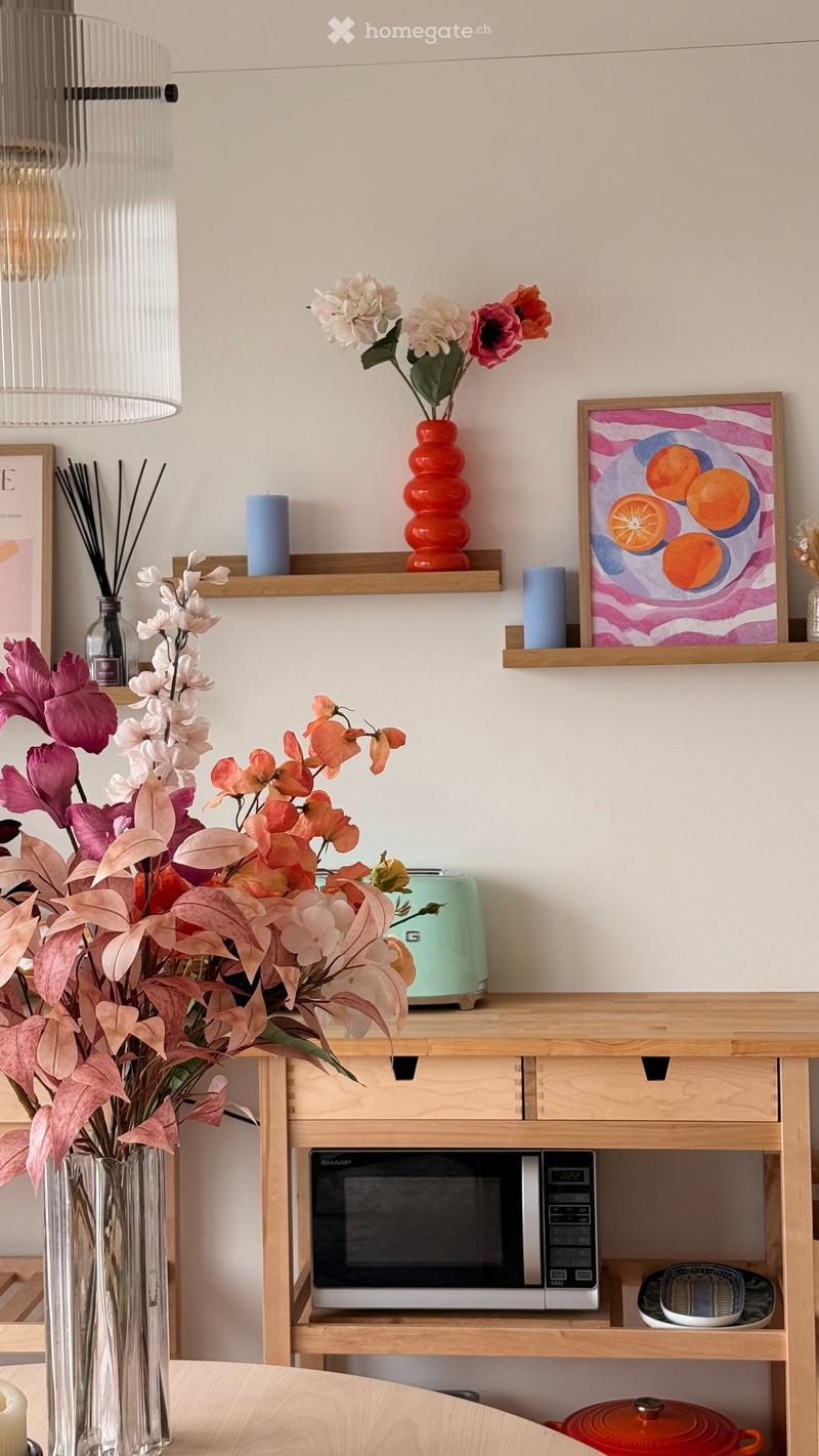 A modern kitchen with a wooden table, a vase with flowers, a microwave, a toaster, and decorative items on shelves.
