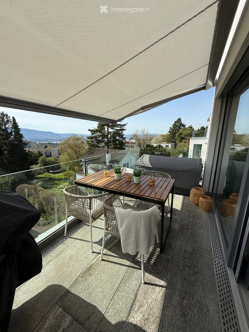 Balcony with a wooden table, two wicker chairs, a cushion, and small flower pots, offering a scenic view of the surrounding area.