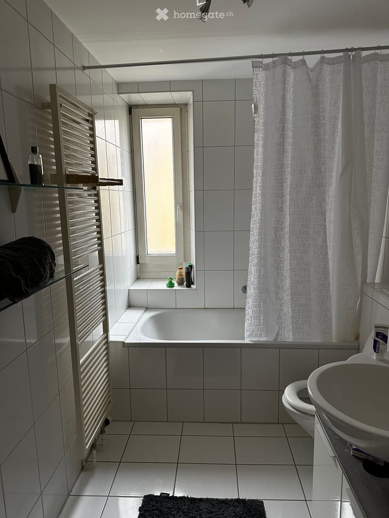 Modern bathroom with white tiles, bathtub, toilet, sink, radiator, and a shower curtain featuring city names. A small window above the bathtub allows natural light.