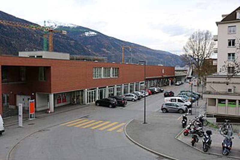 Modern commercial building with a flat roof and large windows, surrounded by a parking lot with several cars and motorcycles.