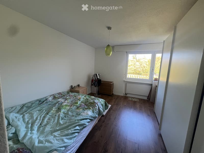 Bedroom with wooden flooring, a bed with a patterned duvet, a nightstand with a lamp, a radiator, and a wardrobe with hanging items.