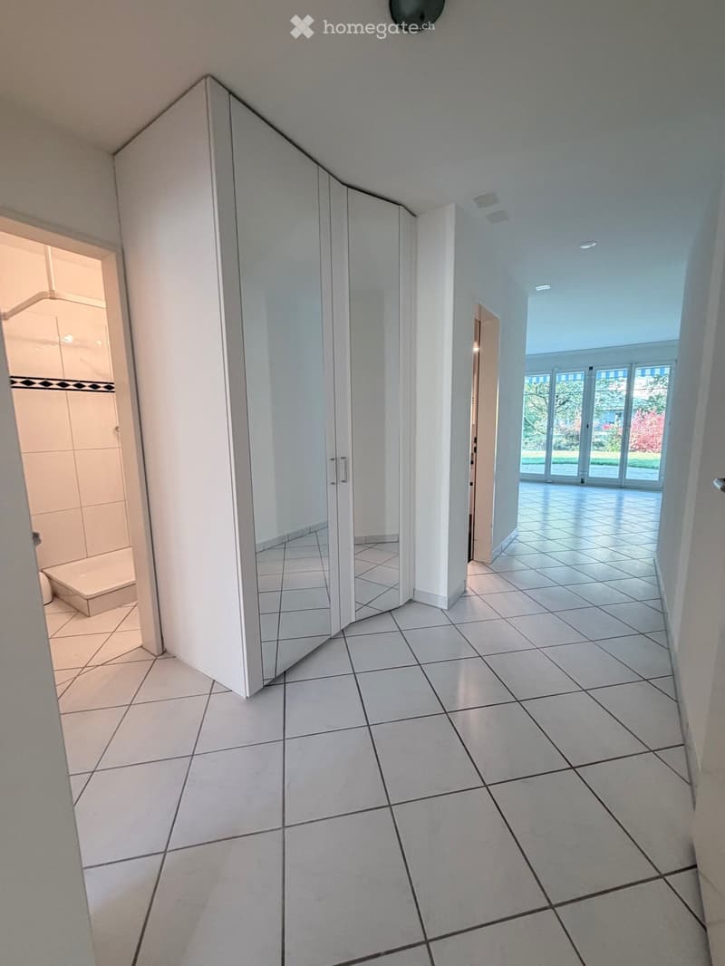 Bright hallway with tiled flooring, white walls, and large mirrored closet doors. Natural light streams in from the right side through large windows, leading to an open space.