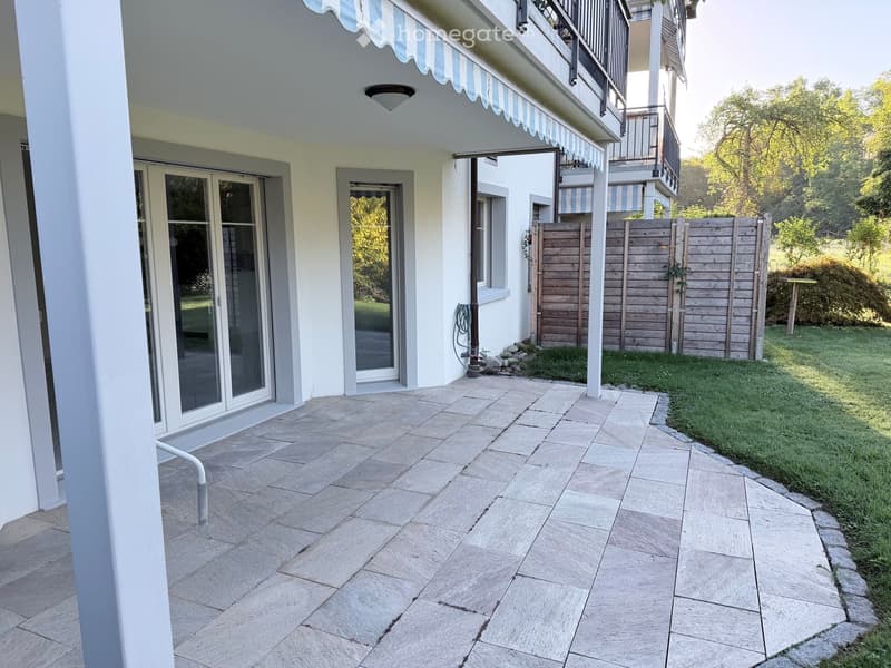 Outdoor patio area with tiled flooring, white walls, glass doors leading to the interior, wooden fence, and greenery in the background.