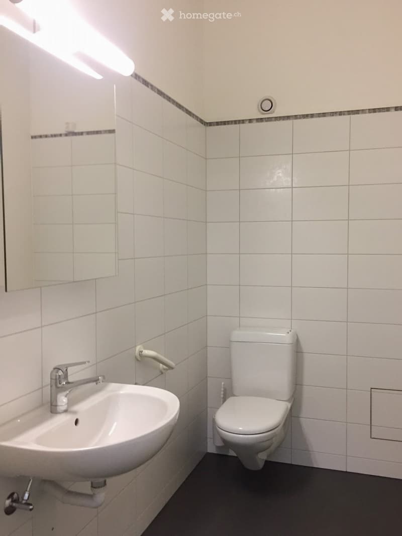 Modern bathroom with white tiles, a sink with a chrome faucet, and a toilet. The walls are tiled with a decorative border, and the floor is dark grey.