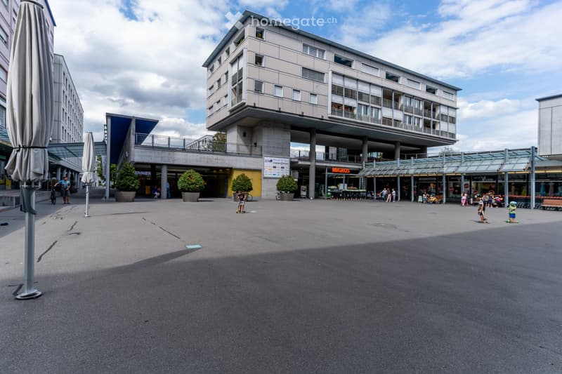 Modern multi-story building with a raised ground floor, large windows, and a covered walkway. The ground floor features shops and seating areas, with people walking and riding scooters.