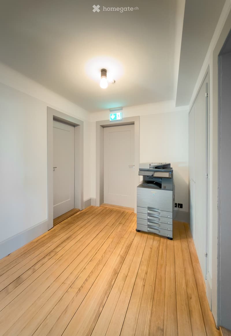 A narrow hallway with wooden flooring, white walls, and two doors. A printer is placed against the right wall near the exit sign.
