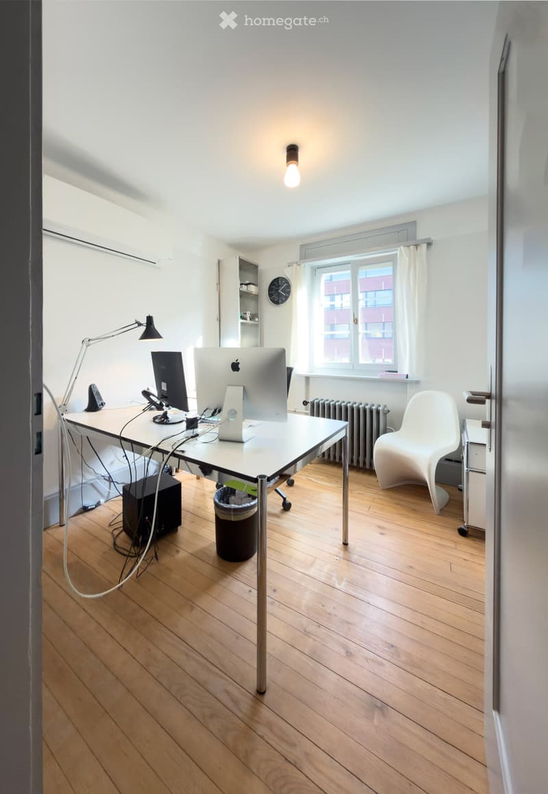 A modern, well-lit room with wooden flooring, a white desk with a computer setup, a white chair, a wall-mounted lamp, a clock, a shelf, and a radiator near the window.